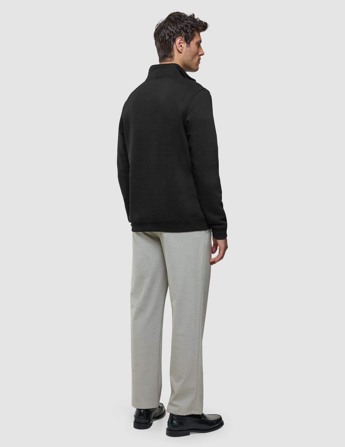 Ein Mann, der ein schwarzes Sweatshirt mit halbem Reißverschluss, eine hellgraue Hose und schwarze Slipper trägt, von hinten gesehen.